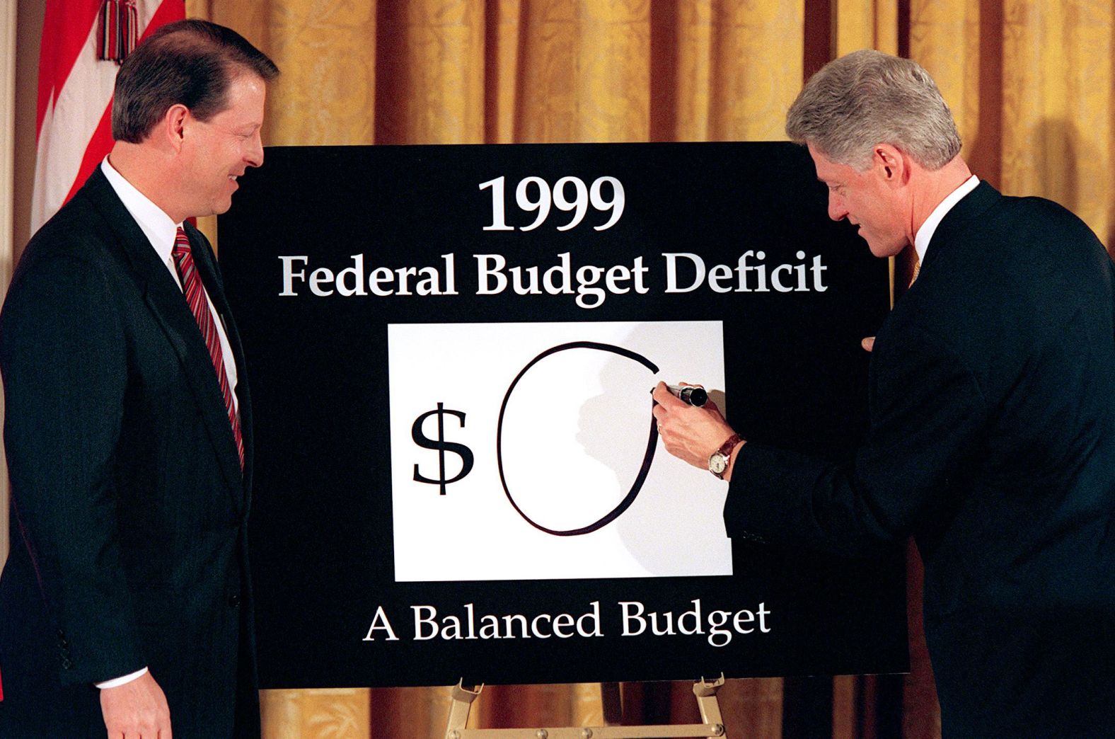 Vice President Al Gore looks on as Clinton writes a "0" on the board, showing what the federal deficit would be after unveiling his balanced budget plan for 1999. The President declared an end to "an era of exploding deficits" as he sent a $1.73 trillion budget to Congress that promised the first surplus in more than three decades.