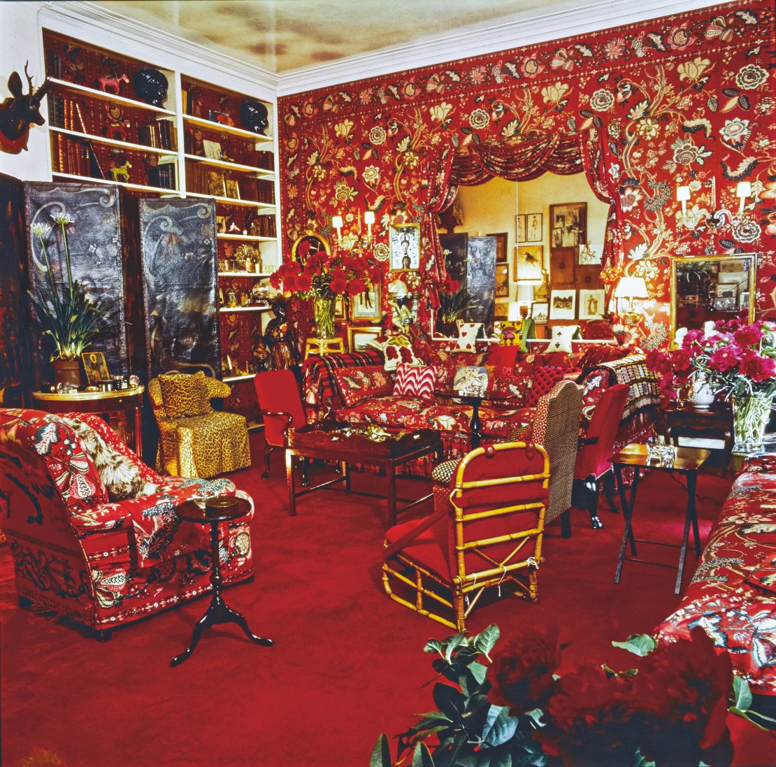 Living room in the Manhattan apartment of fashion editor Diana Vreeland, designed by Billy Baldwin, with dramatic red floral fabric on the walls and as upholstery on the furniture. A painted leather screen rests in front of the bookshelves and there are multiple throw pillows on the couch and a chair covered in leopard.
