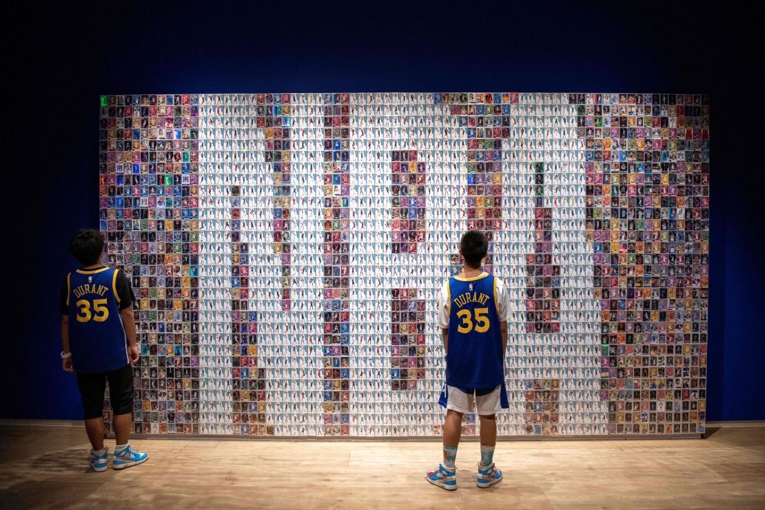 Two Chinese youths stand in front of a wall displaying basketball trading cards at the NBA exhibition in Beijing on August 19, 2019.