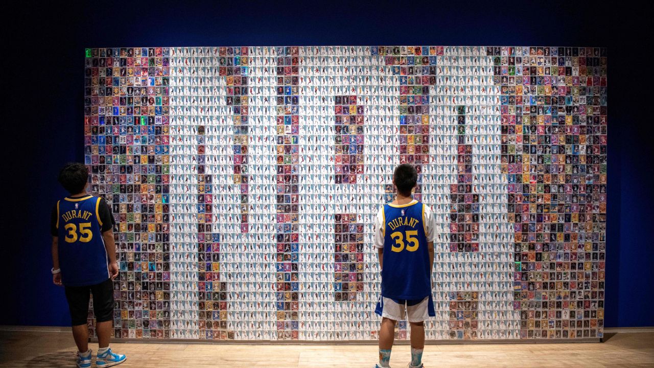 Two Chinese youths stand in front of a wall displaying basketball trading cards at the NBA exhibition in Beijing on August 19, 2019.
