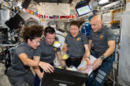 Four Expedition 61 crewmembers unpack fruit and other space-friendly food from a storage bag.