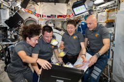 Four Expedition 61 crewmembers unpack fruit and other space-friendly food from a storage bag.