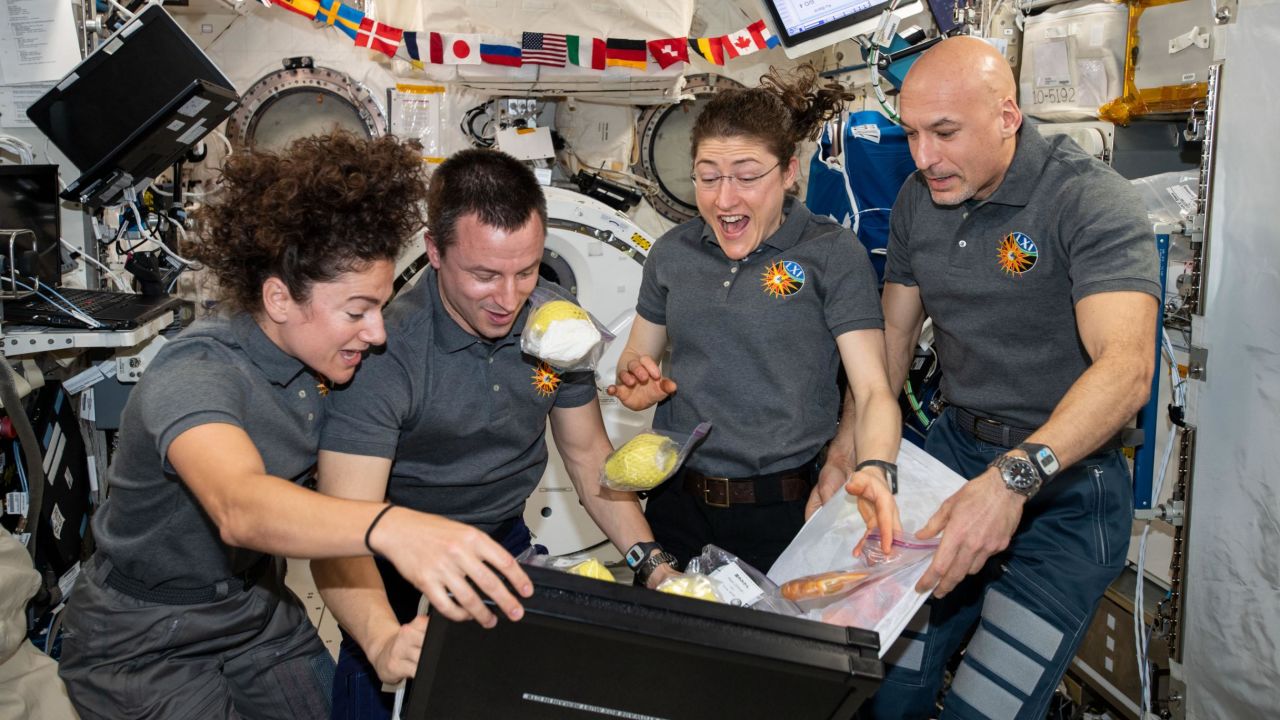 Four Expedition 61 crewmembers unpack fruit and other space-friendly food from a storage bag.