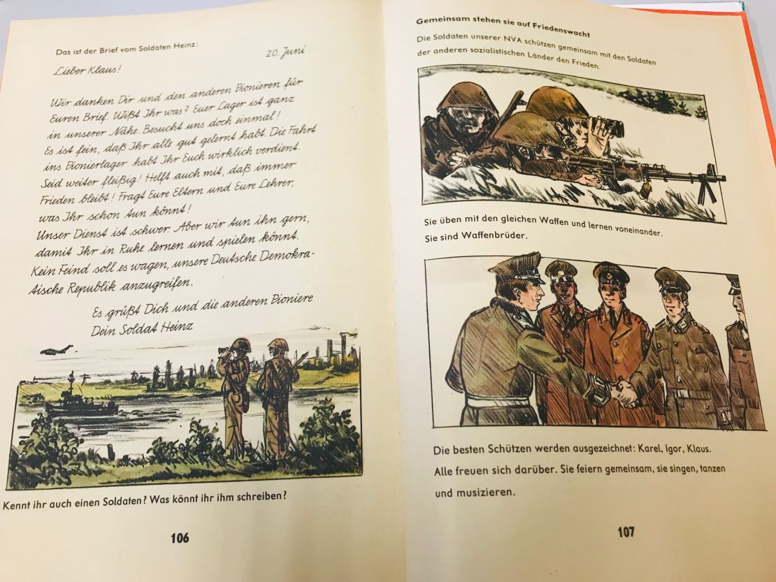 A first grade textbook features an imaginary letter from 'Soldier Heinz' to a pupil. It reads: "Our army service is difficult. But we keep on doing it so you can study and play in peace. No enemy should dare to attack our German Democratic Republic."