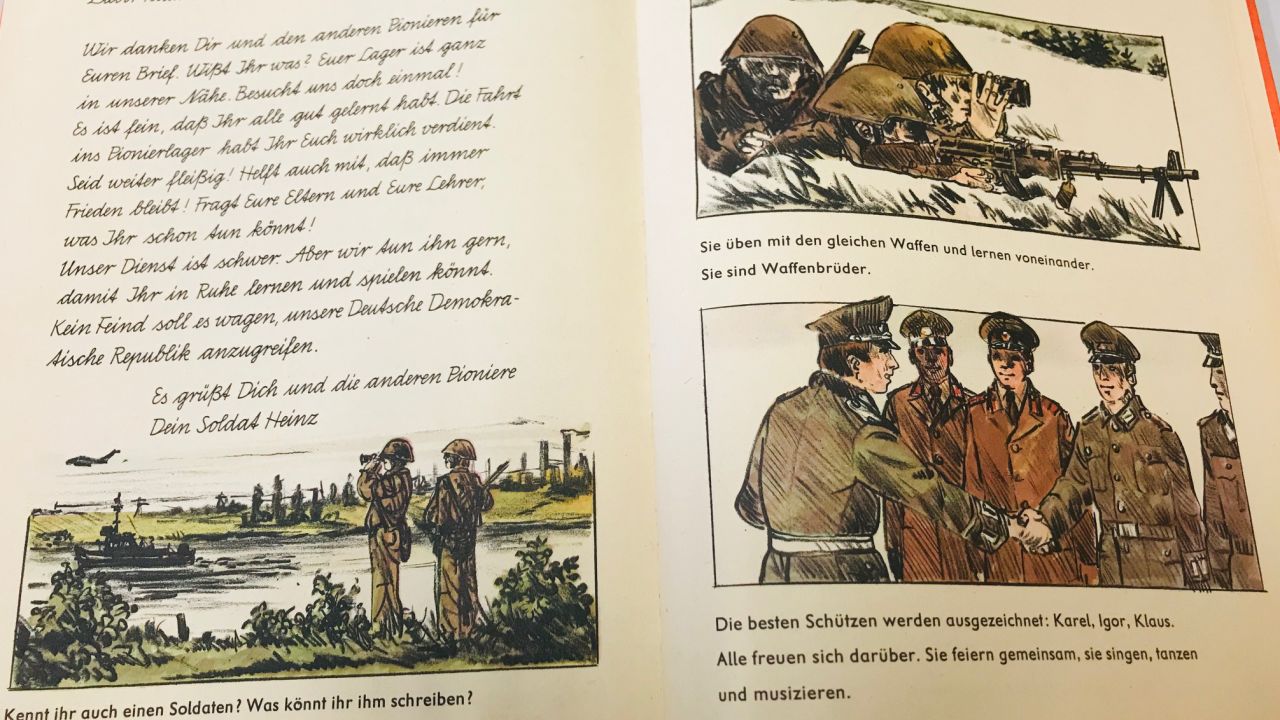 A first grade textbook features an imaginary letter from 'Soldier Heinz' to a pupil. It reads: "Our army service is difficult. But we keep on doing it so you can study and play in peace. No enemy should dare to attack our German Democratic Republic."