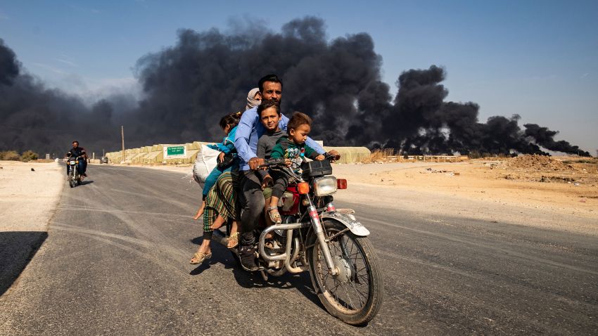 Displaced people, fleeing from the countryside of the Syrian Kurdish town of Ras al-Ain along the border with Turkey, ride a motorcycle together along a road on the outskirts of the nearby town of Tal Tamr on October 16, 2019 as they flee a deadly cross-border Turkish offensive that has sparked an international outcry, with smoke plumes of tire fires billowing in the background to decrease visibility for Turkish warplanes in the area. (Photo by Delil SOULEIMAN / AFP) (Photo by DELIL SOULEIMAN/AFP via Getty Images)