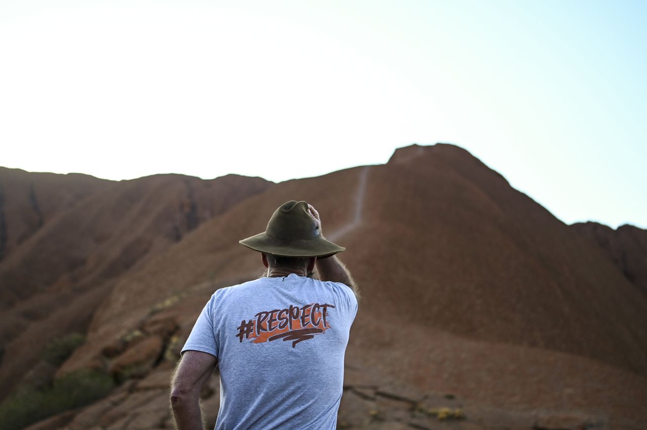 A man wearing a t-shirt saying 'I chose not to climb' stands next to tourists lining up to summit Uluru on October 25.