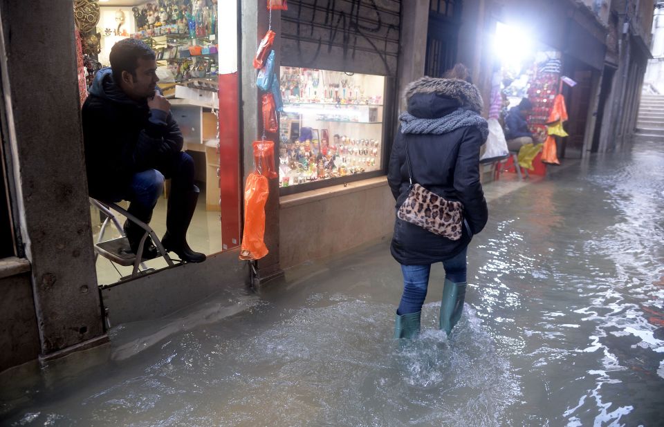 A man sits on a stepladder inside a shop as a woman walks down a flooded street.