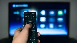 Hand of man pointing remote control at working television screen.