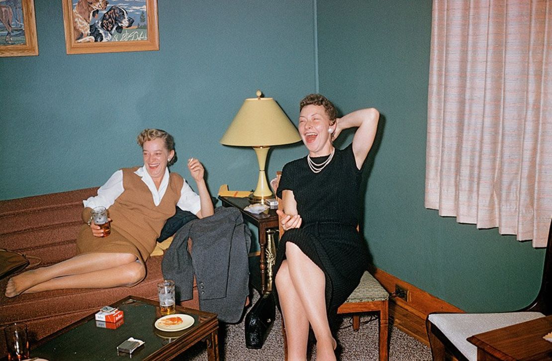 The photo shows two women sitting in someone's teal-colored living room, drinking beer, smoking cigarettes and laughing uproariously at something off-camera.