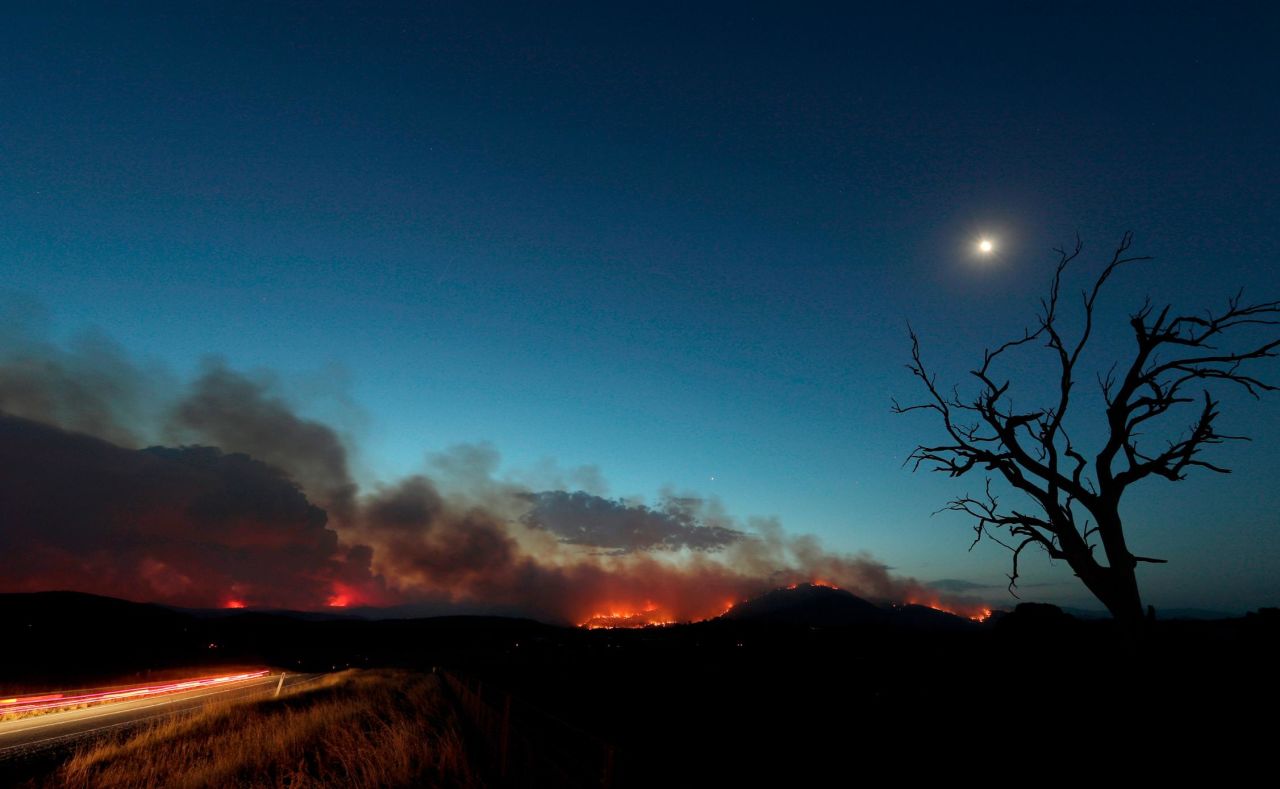 In this long-exposure photo, a car's taillights streak at left as a wildfire glows at dusk near Clear Range, Australia, on Friday, January 31.