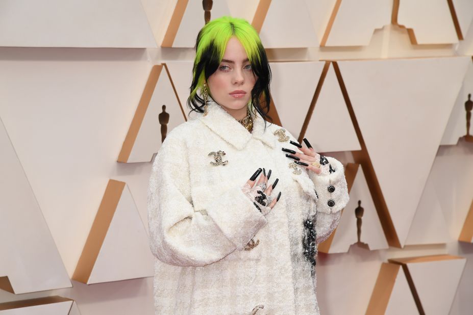 Billie Eilish walks the red carpet in a white-on-white houndstooth set, with a jacket featuring the Chanel's interlocking "C" logo. The singer is also sporting lace fingerless gloves with layered rings and necklaces, crystal accents on her nails and a messy hairdo in her signature two-tone color palette of lime green and black.