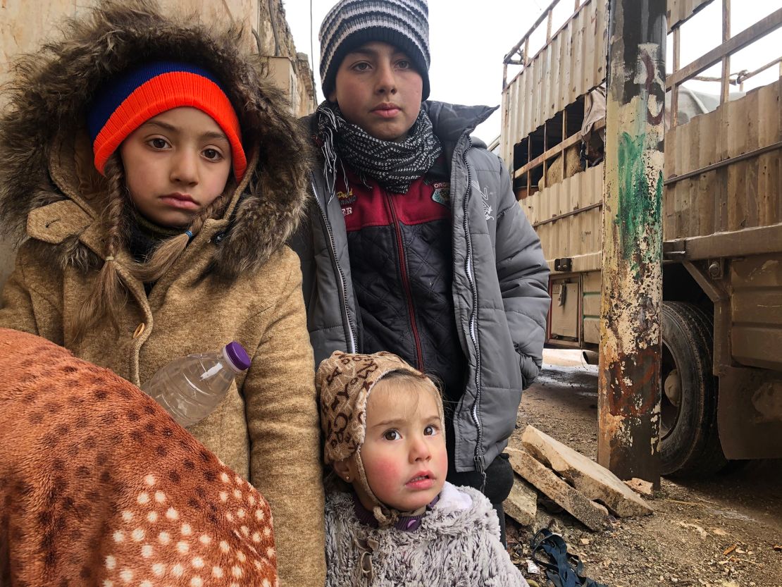 The kids wait patiently and without complaint outside their house in the last opposition-held territory in Syria, as their mother packs their belonging in the back of a truck.
