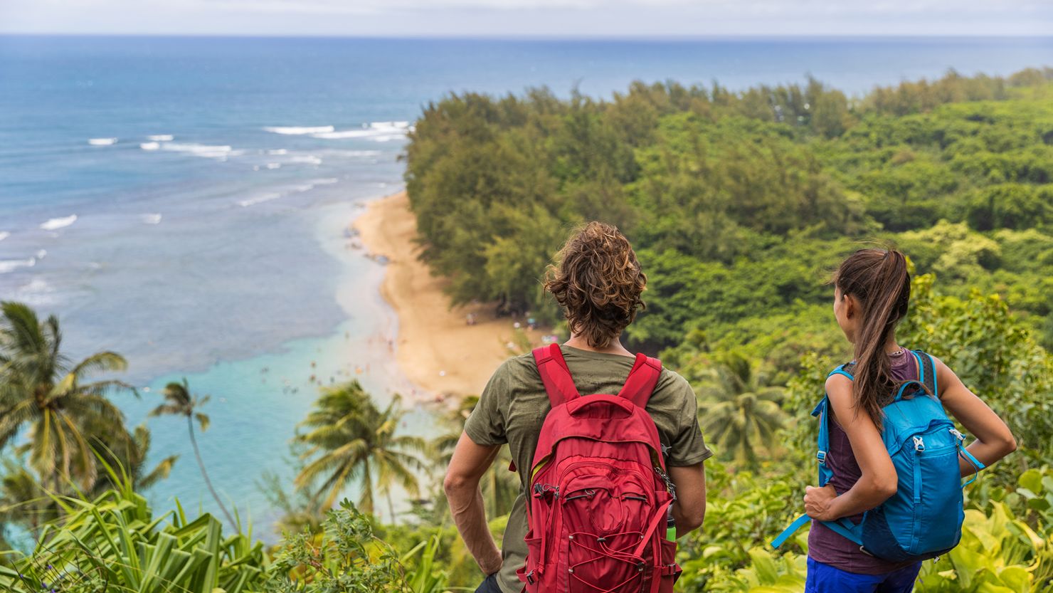 Hikers couple hiking on Kalalau trail overlooking beach coastal walk two tourists with backpacks walking outdoor in Kauai island, Hawaii summer travel leisure activity active lifestyle.