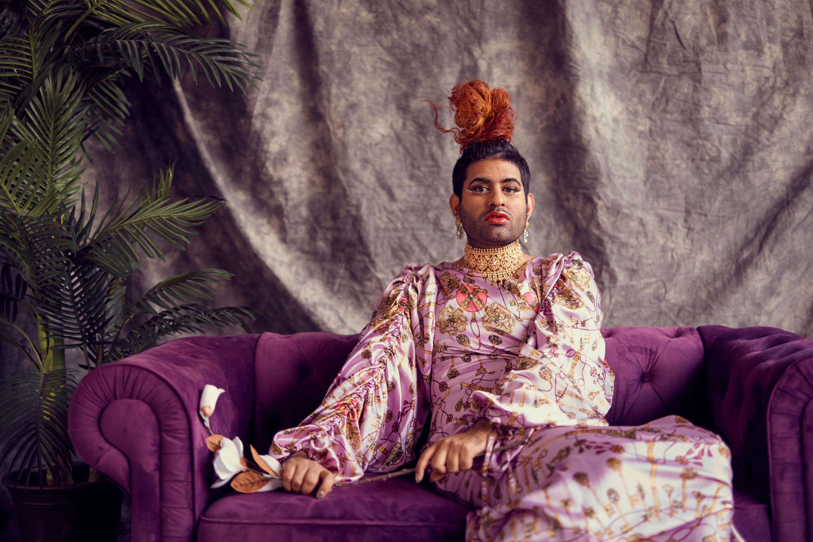 A regal Vaid-Menon sits against a purple couch and luxurious background. They wear a long sleeve high neck dress with gold jewelry.