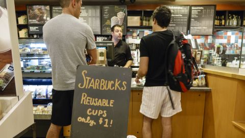 Interior of Starbucks Coffee with customers standing at the counter behind reusable cups sign.
