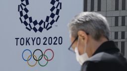 A man wearing a face mask walks before the logo of the Tokyo 2020 Olympic Games displayed on the Tokyo Metropolitan Government building in Tokyo on March 24, 2020. - The International Olympic Committee came under pressure to speed up its decision about postponing the Tokyo Games on March 24 as athletes criticised the four-week deadline and the United States joined calls to delay the competition. (Photo by Kazuhiro Nogi/AFP/Getty Images)