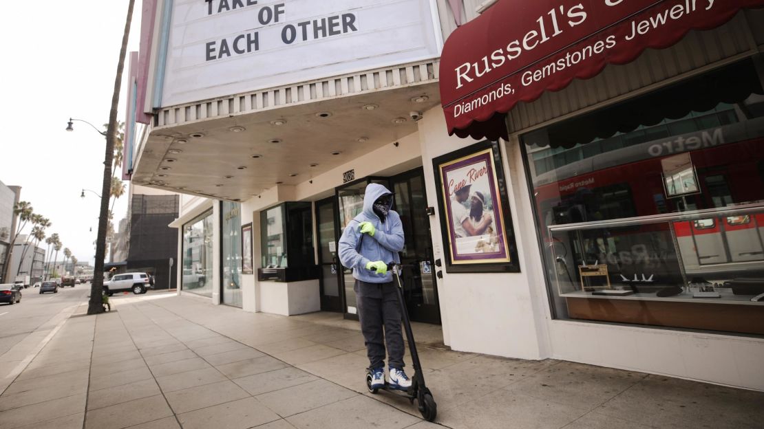 A man wears gloves and a bandana across his face while riding a scooter past a shuttered movie theater on March 18, 2020 in Beverly Hills, California. 