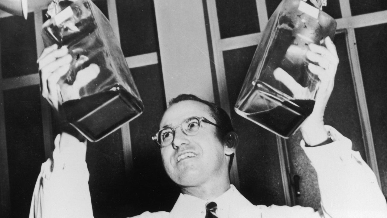 Low-angle view of American scientist and physician Jonas Salk (1914 - 1995), wearing a white lab coat, smiling while holding up two decanters containing the anti-polio vaccine that he developed.  (Photo by Archive Photos/Getty Images)