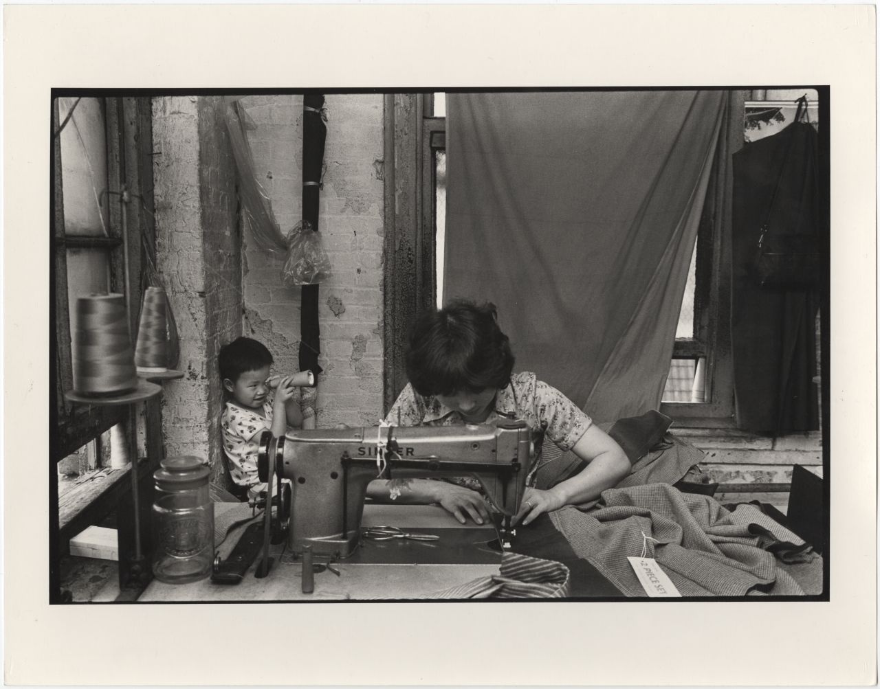 A woman sewing at a garment factory on Canal Street in Manhattan's Chinatown, while a child plays behind her, circa 1984.