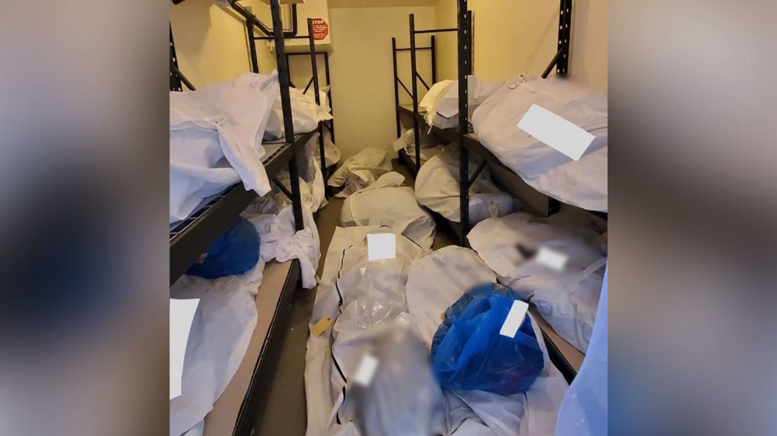 Body bags and blue bags containing the personal effects of the deceased are seen piled inside a portable refrigerator storage unit outside the hospital.