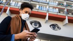 A young woman wearing a protective mask looks at her smartphone while passing by a graffiti representing two big watching eyes in Berlin, Germany on April 1, 2020. Illustrative Editorial (Photo by Emmanuele Contini/NurPhoto via Getty Images)