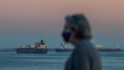 A woman wearing face mask walks on the ocean front while Oil tankers are seen anchored off the coast of Long Beach, California, after sunset on April 25, 2020. - According to a news release issued by the United States Coast Guard, there were 27 tankers off the Southern California coast as of April 23 afternoon. Companies are using the tankers to store excess supplies of crude oil due to lack of demand during the novel coronavirus pandemic, US media reported. (Photo by Apu Gomes/AFP/Getty Images)