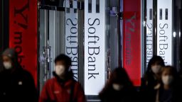 Pedestrians wearing protective masks walk past illuminated signage for SoftBank Corp. and the company's Y!mobile brand displayed outside a store at night in Tokyo, Japan, on Tuesday, April 14, 2020.