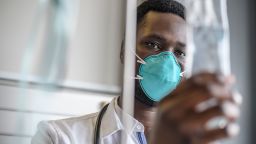 Personal perspective close-up of young African male medical professional wearing lab coat and N95 face mask checking IV fluid bag in hospital room.