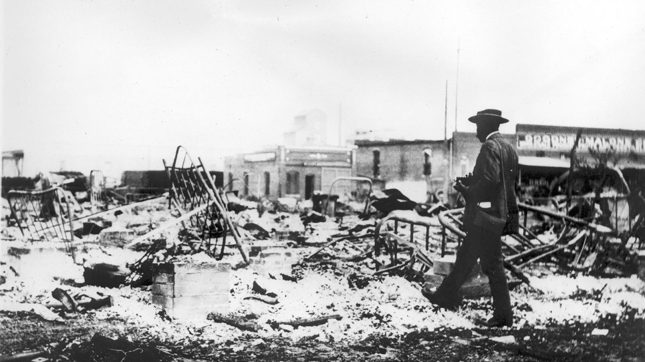 An African-American man with a camera looking at the skeletons of iron beds which rise above the ashes of a burned-out block after the Tulsa Race Riot, 1921. 