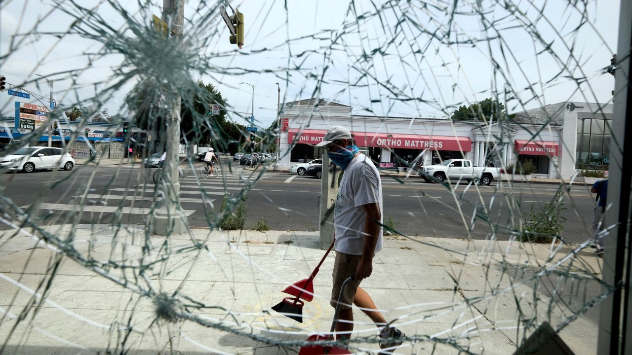 A volunteer sweeps up broken glass behind a shattered store glass door, Sunday, May 31, 2020, in Los Angeles, following a night of unrest and protests.
