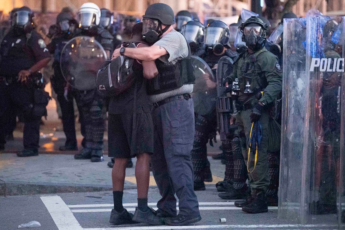 An officer clad in a helmet and mask hugs a protester.