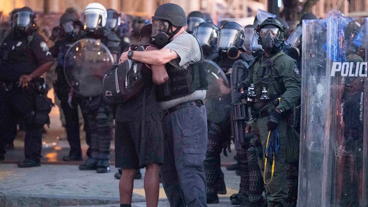 An officer clad in a helmet and mask hugs a protester.