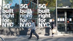 People wearing protective masks and gloves pass by a Verizon store advertising 5G in New York amid the coronavirus pandemic on April 28, 2020 in New York City, United States. COVID-19 has spread to most countries around the world, claiming over 217,000 lives with over 3.1 million cases. (Photo by Alexi Rosenfeld/Getty Images)