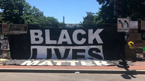 A large Black Lives Matter flag hangs from the fence, with the Washington Monument spotted in the background. 