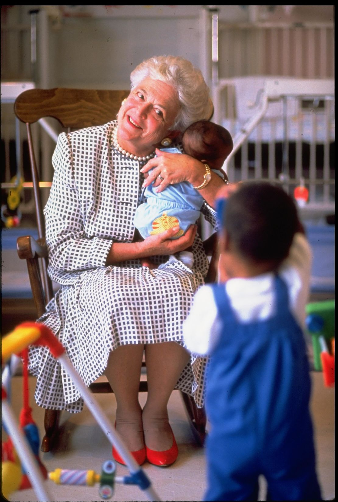 First Lady Barbara Bush holding baby while two-year-old child takes a photo wtih a toy camera at hospice for children with  AIDS. 