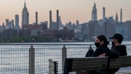 NEW YORK, NEW YORK - MAY 21: A couple wearing masks sit on a bench watching the sunset over the Manhattan skyline in Domino Park, Brooklyn amid the coronavirus pandemic on May 21, 2020 in New York City. COVID-19 has spread to most countries around the world, claiming over 334,000 lives with over 5.1 million cases. (Photo by Alexi Rosenfeld/Getty Images)