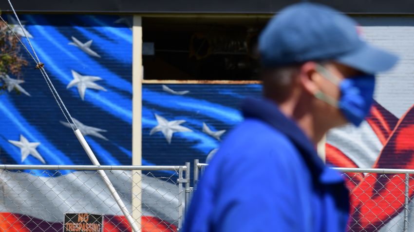 A pedestrian wearing a mask walks across the street from a fenced off building with a large American flag mural displayed on Sunday April 19, 2020 in Arlington, VA. The coronavirus has altered daily life in the region and the country. (Photo by Matt McClain/The Washington Post via Getty Images)
