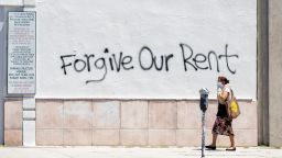 A woman wearing a mask walks past a wall bearing a graffiti asking for rent forgiveness on La Brea Ave on National May Day amid the Covid-19 pandemic,  May 1, 2020, in Los Angeles, California. 