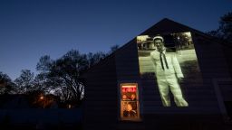 An image of veteran James Mandeville is projected onto the home of his daughter, Laurie Mandeville Beaudette, as she looks out a window with her son, Kyle, left, and husband, Mike, in Springfield, Mass., Tuesday, May 12, 2020. Mandeville, a U.S. Navy veteran and resident of the Soldier's Home in Holyoke, Mass., died from the COVID-19 virus at the age of 83. Seeking to capture moments of private mourning at a time of global isolation, the photographer used a projector to cast large images of veterans on to the homes as their loved ones are struggling to honor them during a lockdown that has sidelined many funeral traditions. (AP Photo/David Goldman)
