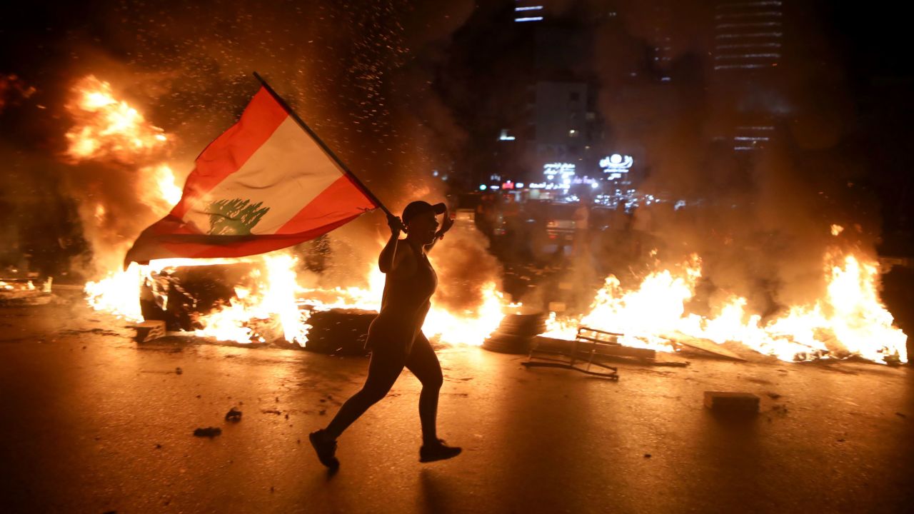 TOPSHOT - A protester holding the Lebanese flag runs as protesters block the Jounieh Tripoli highway with flaming tires set aflame during a demonstration against dire economic conditions in Jal el Dib North East of the Lebanese capital Beirut late on June 11, 2020. - The Lebanese pound sank to a record low on the black market on June 11 despite the authorities' attempts to halt the plunge of the crisis-hit country's currency, money changers said. Lebanon is in the grips of its worst economic turmoil in decades, and holding talks with the International Monetary Fund towards securing billions in aid to help overcome it. (Photo by Patrick BAZ / AFP) (Photo by PATRICK BAZ/AFP via Getty Images)