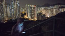 A man wearing a protective mask is illuminated by a smartphone as residential buildings stand in the background at night in Hong Kong, China, on Thursday, Feb. 20, 2020.