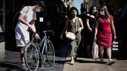 A man wearing a protective mask unlocks his bicycle as women walk holding shopping bags in a street, in Bordeaux, on July 18, 2020. (Photo by Philippe LOPEZ / AFP) (Photo by PHILIPPE LOPEZ/AFP via Getty Images)
