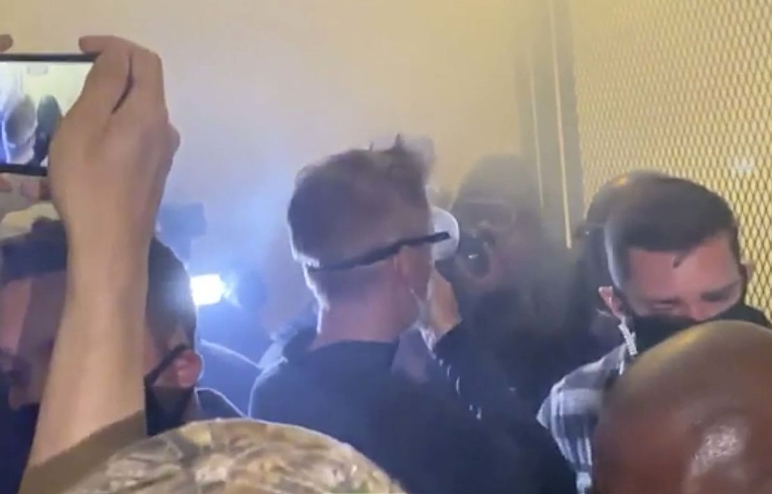 In this image made from video released by Jonathan Maus, Portland Mayor Ted Wheeler, center in black with goggles looking away, stands at a fence guarding a federal courthouse as tear gas drifts by early July 23, 2020, in Portland Oregon.