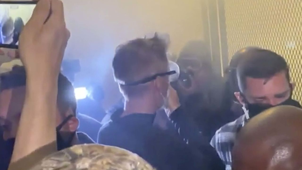 In this image made from video released by Jonathan Maus, Portland Mayor Ted Wheeler, center in black with goggles looking away, stands at a fence guarding a federal courthouse as tear gas drifts by early July 23, 2020, in Portland Oregon.