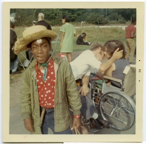 In another vintage color snapshot at "Crip Camp," Cary Walker squints at the camera with a bemused look.  Behind him, a couple sitting in wheelchairs share a romantic embrace. 