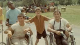 Caption:, 1968 outdoor color snapshot. Four male campers, two in wheelchairs, smile for the camera. Behind them a large group assembles in the Jened field., Visual Description:, two sitting in manual wheelchairs and two standing, smile for the camera. The boy standing in the middle has his arms on the backs of the wheelchairs to either side of him. Behind them a large group assembles in the Camp Jened field., Alt Text:, Vintage snapshot of four Jened campers, smiling for the camera.