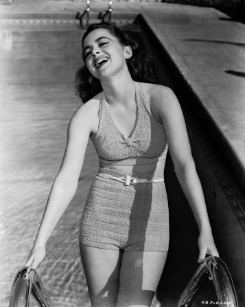 De Havilland dons a two-piece swimsuit and laughs as she ascends from a pool, in a photograph for Warner Brothers (circa 1938).<br />