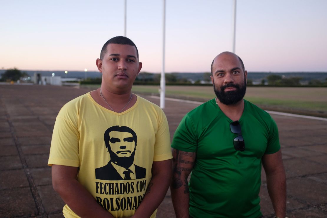 Supporters of President Bolsonaro gather outside his residence. One wears a t-shirt that reads "Sticking with Bolsonaro" on the front and "All power to the people" on the back.