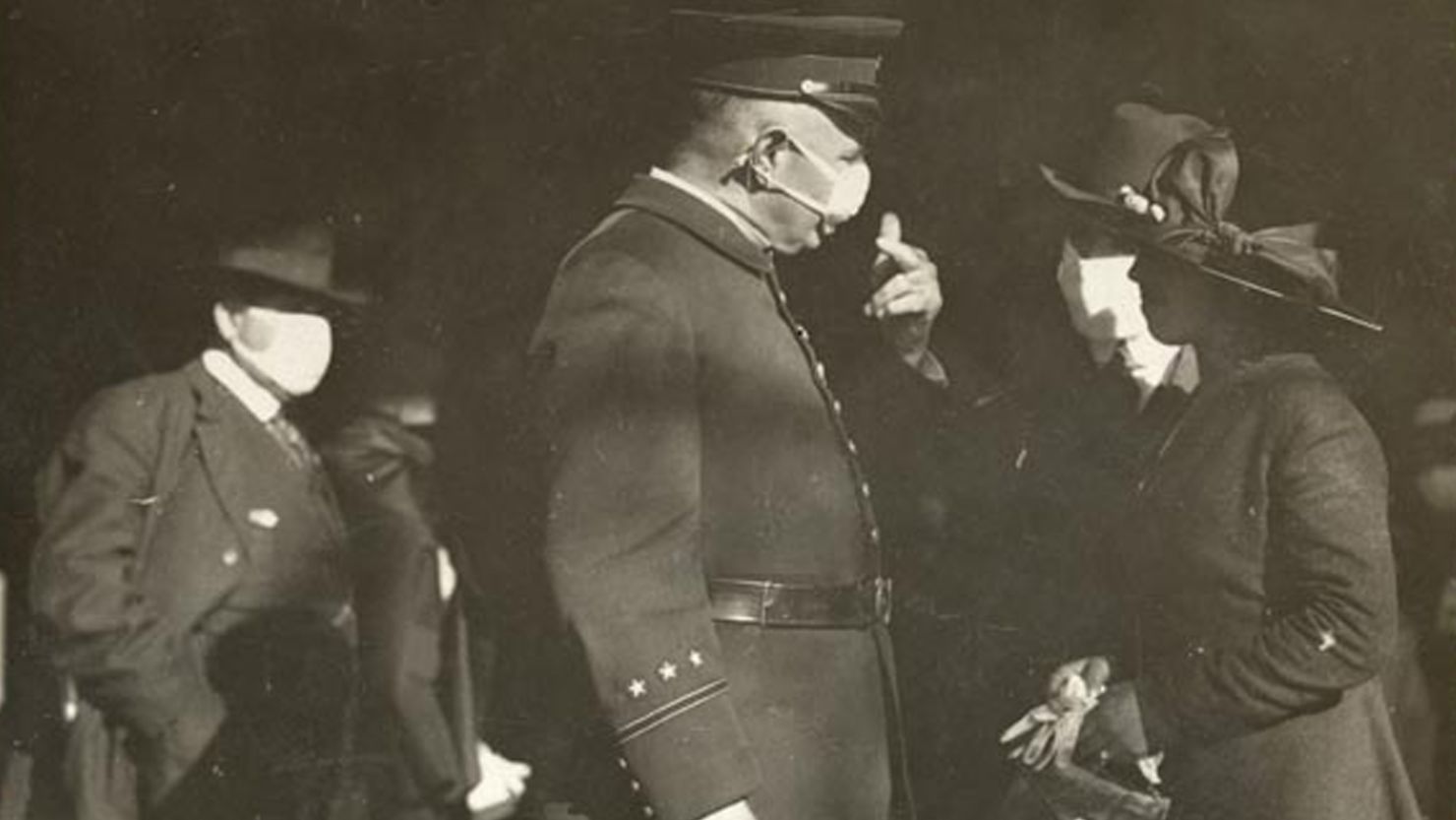 A San Francisco police officer talks to a couple about wearing masks in 1918. The man is wearing a mask but the woman is not, prompting a warning from the officer.